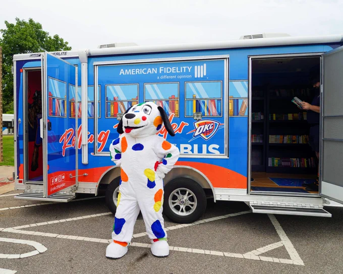 Metro Library's colorful spotted dog mascot, Spoticus, standing in front of the blue and orange Rolling Thunder Book Bus with the doors open.
