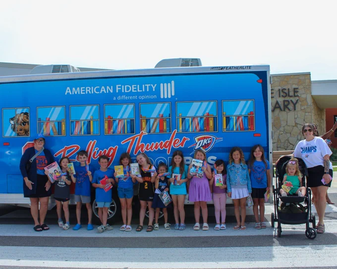 Group of children and two adults standing in front of a blue Rolling Thunder Book Bus holding books outside the Belle Isle Library.