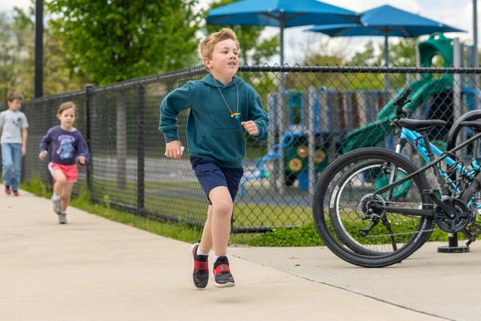 A group of children runs energetically along a sidewalk near a playground. The mood is lively and playful.