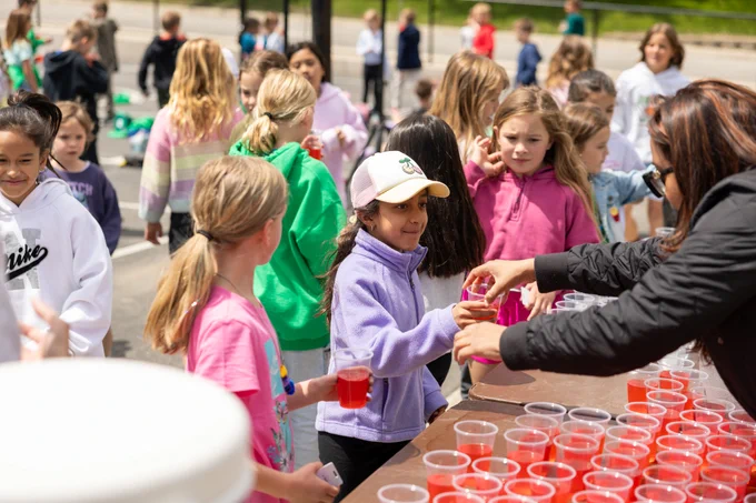 A group of children gather outdoors, receiving red drinks from a table. The event appears lively and social, with children dressed in colorful casual clothing. The mood is cheerful and communal.