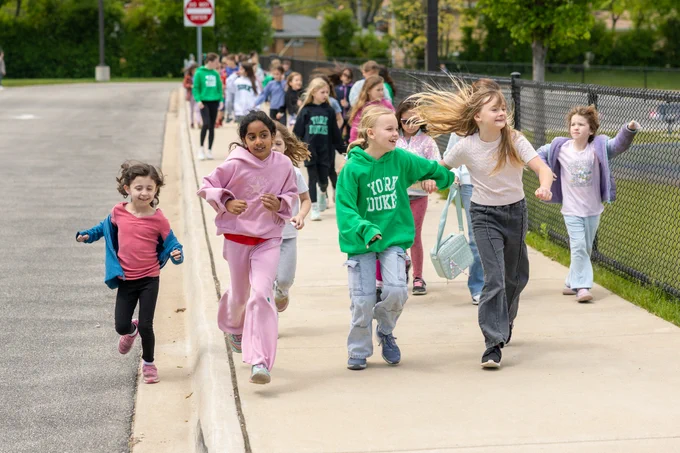 A group of children joyfully running along a sidewalk near a fence. They appear energetic and happy, dressed in casual clothing.