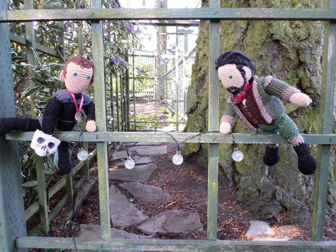 Crochet dolls of Karl Tanner & Robert Rogers hanging a section of the old solar lights on the lattice fence for the squirrels to have something to chew on. Yard in background. 