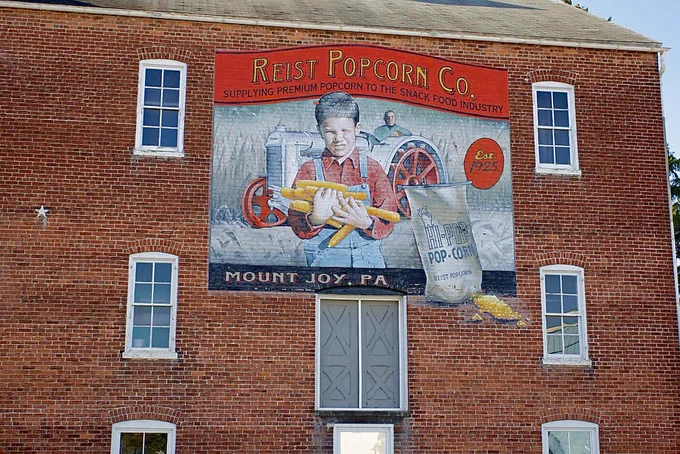 A large mural on the side of a historic brick building depicting a young farmer boy holding ears of corn with a man riding a tractor behind him and the red banner with Reist Popcorn Company above the scene