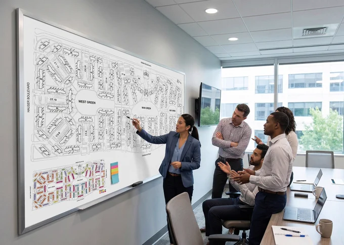 A businesswoman marks up a whiteboard as four professionals in business casual attire interact with her about the diagram organizational diagram she's illustrating
