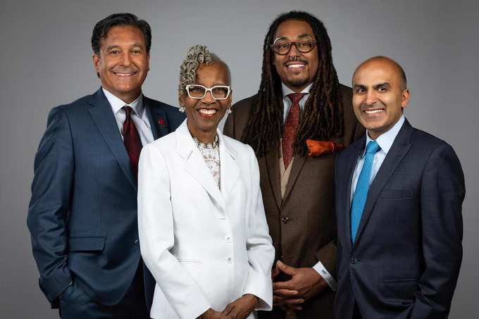 Four professionals in formal business attire pose side by side against a neutral studio background, including one person in a white suit at center and others in dark suits and ties.
