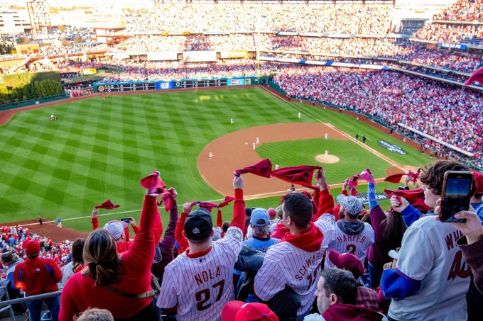 Fans in Phillies jerseys and caps cheer enthusiastically at a packed baseball stadium during a daytime game.
