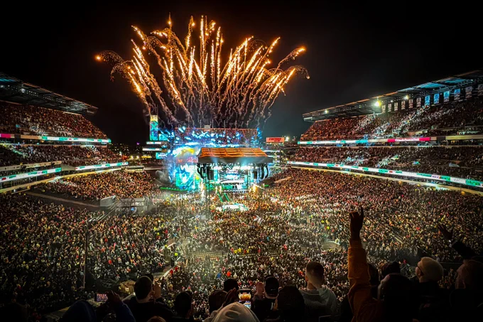 Fireworks explode above a packed stadium during a nighttime concert with a brightly lit stage and cheering crowd.