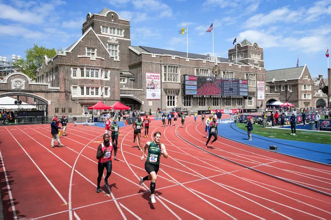 Runners competing on a red track in front of a large historic brick building under a blue sky.