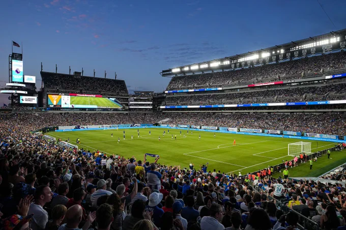 A packed stadium under evening lights with players actively competing on a vibrant green soccer field.