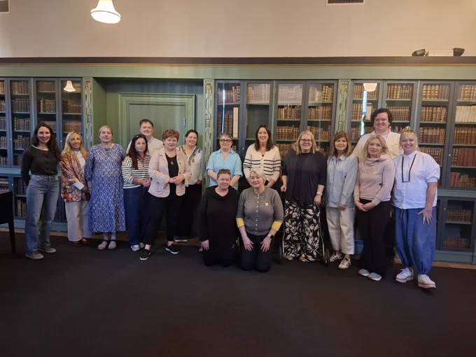 Group of fourteen people posing in front of tall bookshelves filled with old books in a library setting, with two individuals kneeling in front.