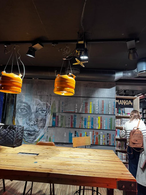 Cozy library corner with wooden table, hanging orange lamps, and a large mural of a face on the wall behind bookshelves filled with colorful books.