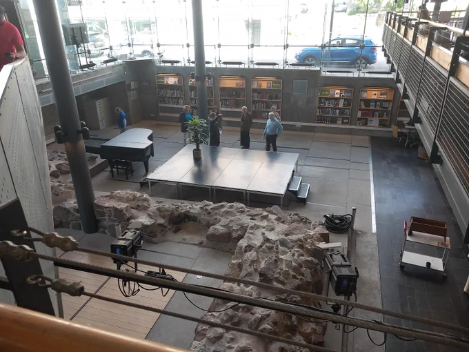 Modern library interior with exposed ancient stone ruins, a small stage, and visitors exploring the space.