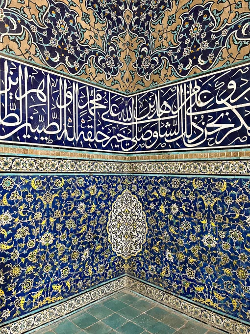 Close-up, vertical perspective looking into a tiled corner of the historic Sheikh Lotfollah Mosque in Isfahan, Iran. The lower half of the walls features dense, intricate blue, yellow, and green floral mosaics meeting at the corner seam. Above this floral pattern, a prominent, wide horizontal band of deep-blue tiles features flowing, elegant white Arabic calligraphy. The upper section transitions into elaborate, swirling arabesque tilework in shades of cream, blue, and brown, meeting a green-tiled floor below.