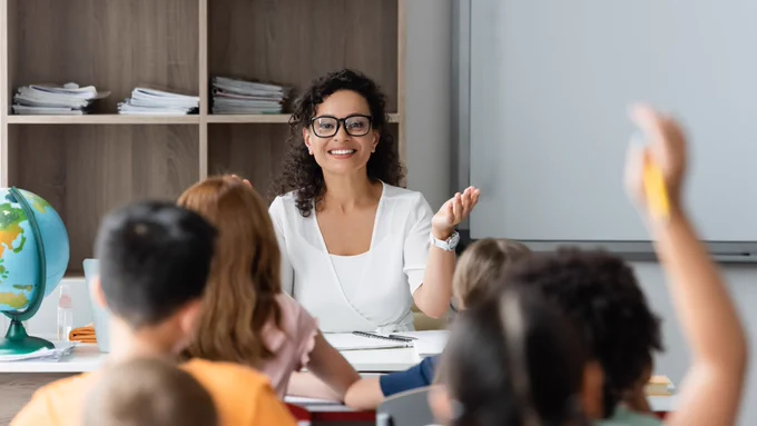 A teacher smiling while leading a classroom discussion, with students seated at desks, one raising their hand.