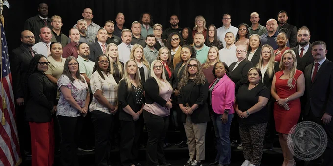 A group of 50 men and women pose for a group portrait. A US flag in seen on the left and the BOP seal placed in the lower-right corner.