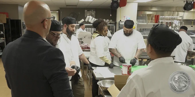 Group of individuals in in a kitchen wearing kitchen uniforms preparing food and being overseen by a man in a suit.