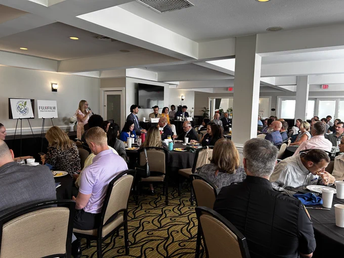 Group of people sitting down listening to a speaker talk. A group of panelists are sitting at the front of the room. People are eating breakfast food inside.