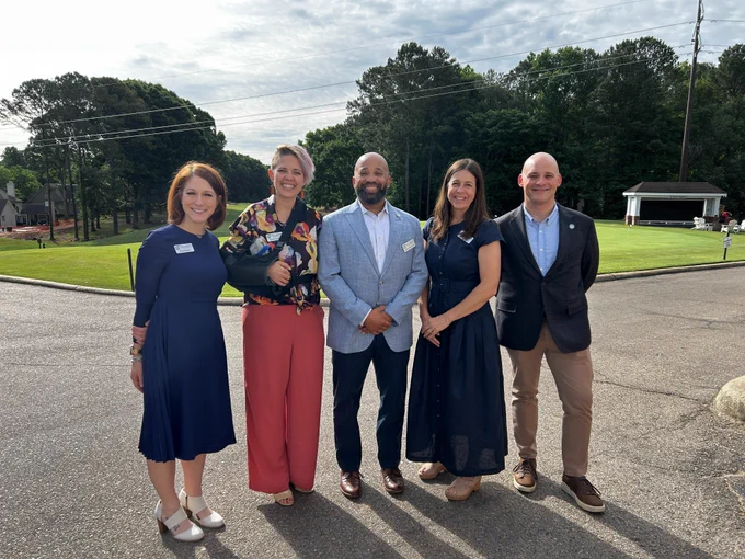 Five people standing in business attire outside smiling at the camera on a sunny day.