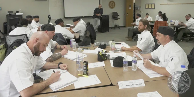 Group of individuals in kitchen uniforms taking notes during a lecture.