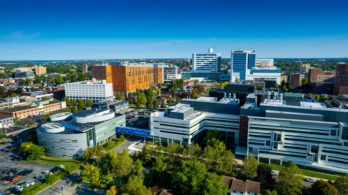 Aerial view of a vibrant urban campus under a clear blue sky. Modern buildings include a large orange structure, a white hospital labeled and a grey circular building. A blue pedestrian bridge reads \"innovation starts here.\"