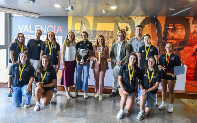 Equipo femenino con medallas y trofeo posando con autoridades frente a un mural que dice VALENCIA.