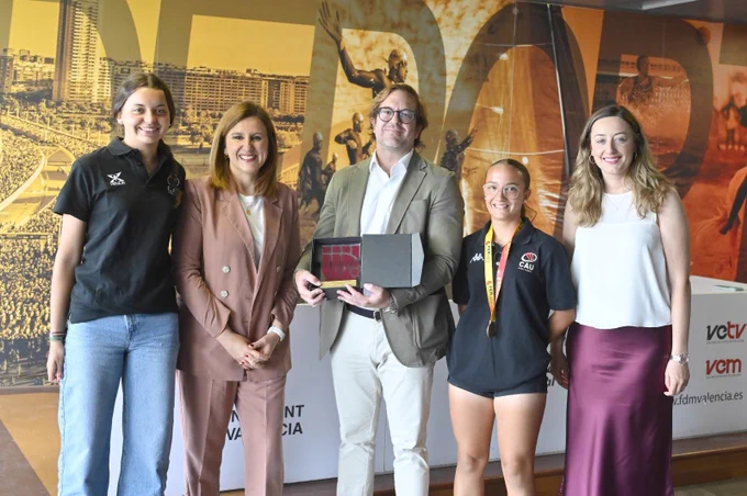 Cinco personas sonrientes ante un mural; hombre sostiene estuche con trofeo; dos jóvenes con ropa deportiva y medalla