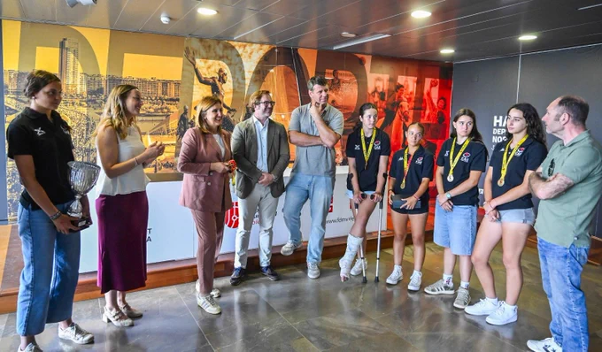 Equipo femenino joven con medallas y trofeo junto a autoridades en recepción; una atleta con yeso y muletas.