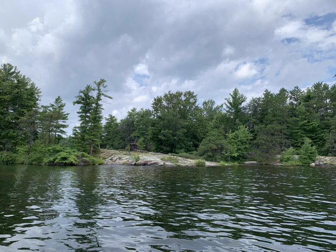 A scenic view of a calm lake with a rocky shoreline in the foreground. Lush green trees line the shore, and a partially cloudy sky is visible above, creating a peaceful natural setting.