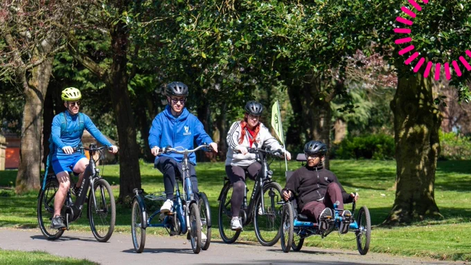 Four people are cycling on a path in a park. They are using a range of adapted cycles. It is a sunny day and there are trees and grass in the background.