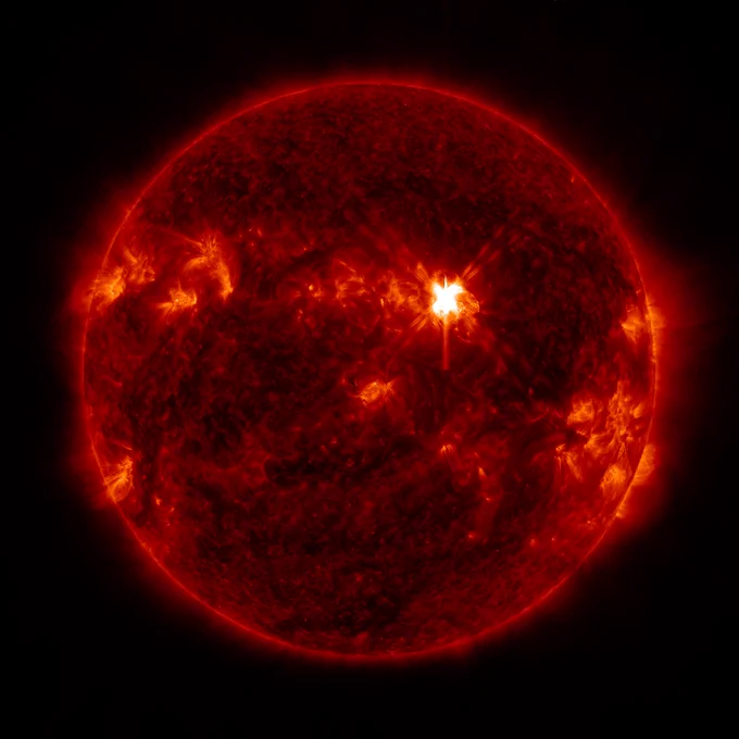 The Sun, shown against the black backdrop of space. The Sun is colorized in red, with dark freckles spotting it and some brighter, more orange areas representing active regions. Toward the upper right, there is a bright white region, showing a flash shaped like an X. This is the solar flare.