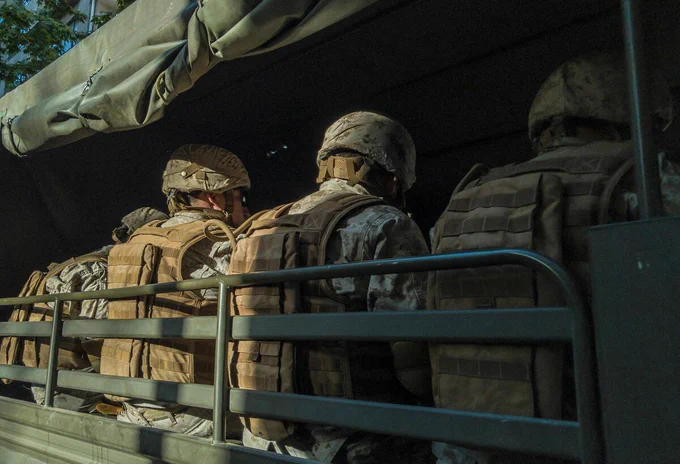 Soldiers in camouflage uniforms and helmets sitting in the back of a military truck, facing forward. The scene is outdoors with sunlight filtering in.

Photo credit: Diego González via Unsplash