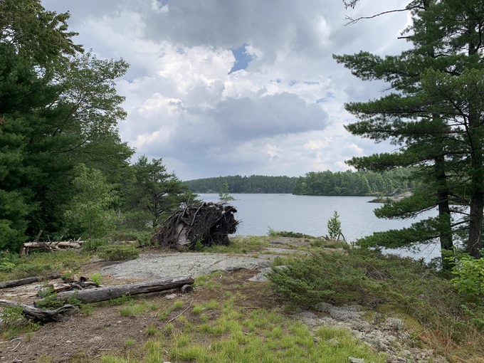 A serene lakeside scene features a calm body of water surrounded by dense green forests and some rocky terrain. A cloudy sky looms above, creating a peaceful atmosphere.