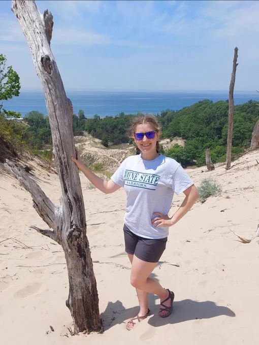 Wayne State University student smiles next to tree with sand, trees, and a body of water in the background