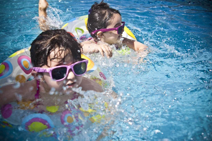 Two children wearing goggles play and splash in a pool while floating on colorful inflatable rings.
