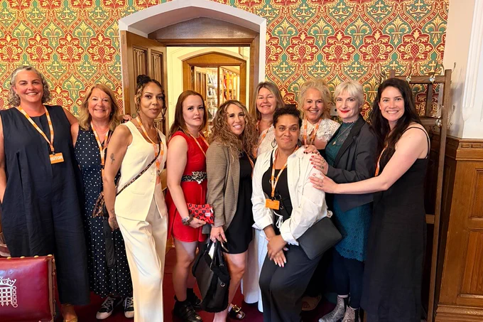 A group of women smile at the camera. They are stood in a line huddled together in a grand room, with wood panelling and patterned wallpaper surrounding a large doorway. In the foreground a red leather chair can be partially seen with the House of Lords logo printed on it.