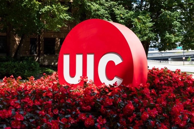 Red UIC sign surrounded by vibrant red flowers and green trees in an outdoor campus setting.