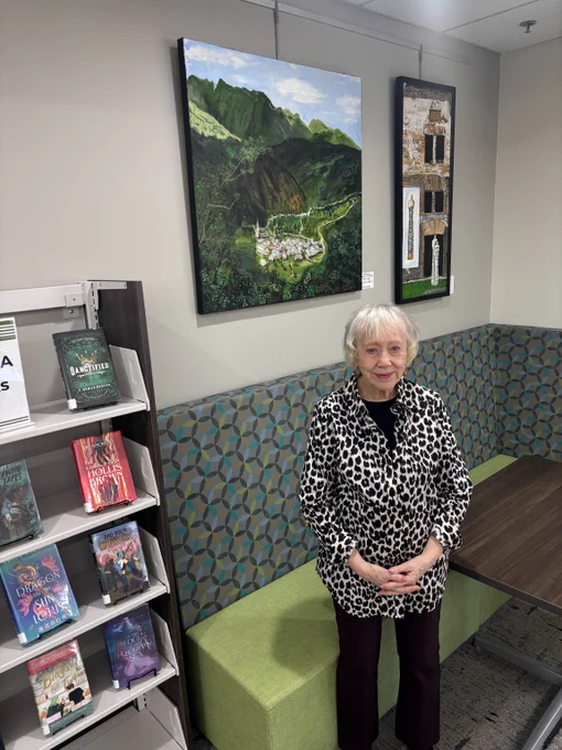 Senior woman stands near two landscape paintings hanging in a corner of a library.