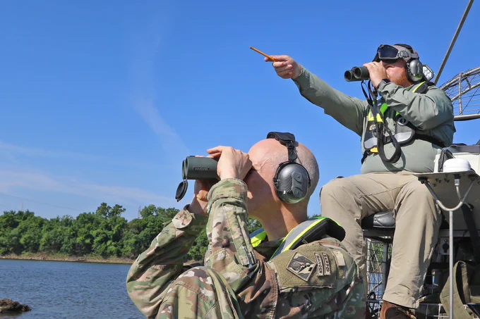 Two men on a boat, hold binoculars to their eyes as they conduct a survey of birds on a lake. 