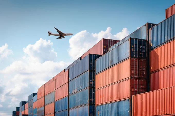 Cargo containers stacked in rows under a blue sky with an airplane flying overhead.