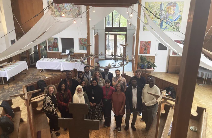 A diverse group of around 20 people pose together for a photo inside a bright, high-ceilinged church hall, with colourful artwork on the walls and a refreshment table in the background.