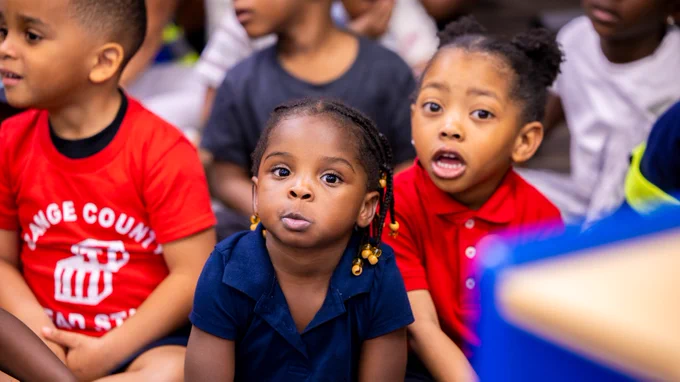 Children sitting together in a classroom, with one wearing a shirt that reads "Orange County Head Start." They appear focused and attentive.