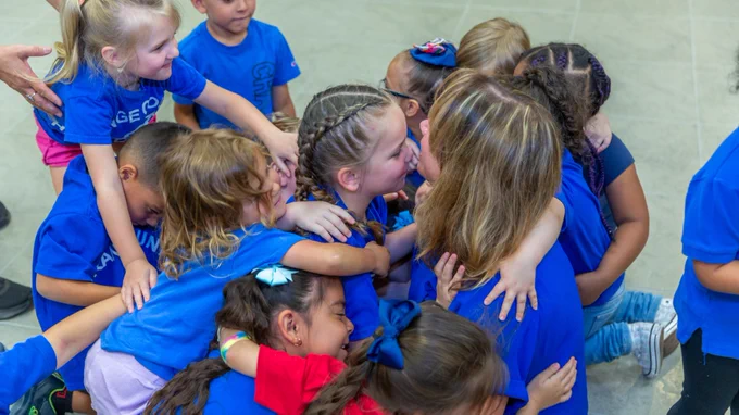 A group of young children in blue shirts are hugging each other enthusiastically in a bright indoor setting.