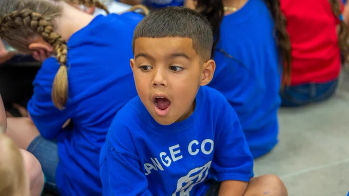 A child wearing a bright blue shirt with "Orange County" printed on it is sitting on the ground, looking to the side with an open mouth expression. Other children in similar outfits are seated nearby.