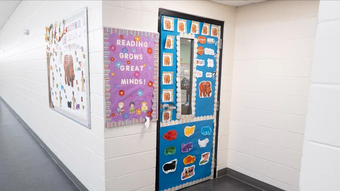A school hallway with white walls. Two colorful bulletin boards are displayed: one on the left featuring a variety of animal images, and another on a door with text reading "Reading Grows Great Minds!" surrounded by images of animals and books.