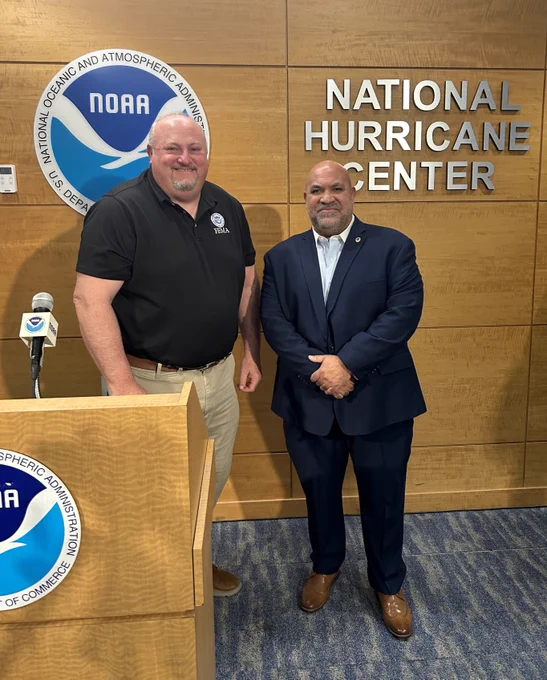 Two men standing in front of the National Hurricane Center sign and NOAA logo on a wooden wall inside an official conference room.