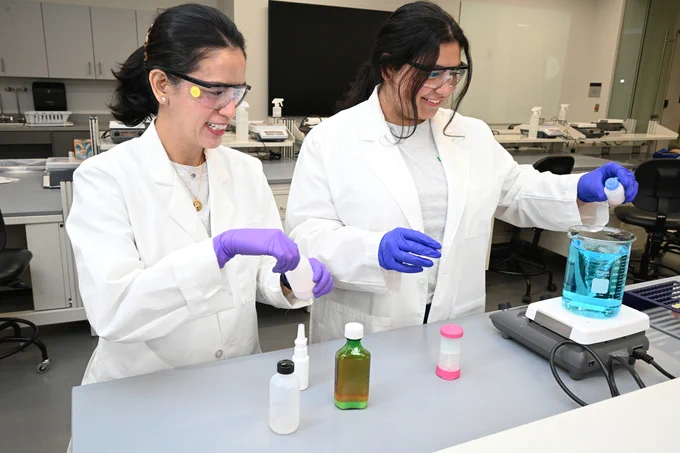 Two individuals in lab coats and gloves are conducting an experiment at a lab bench. They are handling containers with liquid, including a large, transparent beaker filled with a blue liquid on a hot plate stirrer. Various bottles are on the counter. The setting is a modern laboratory with stools and equipment in the background.