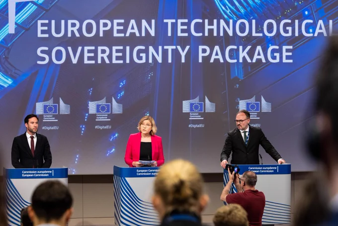 EVP Henna Virkkunen speaks at a European Commission press conference on the European Technological Sovereignty Package, standing at a podium alongside Commissioner Dan Jørgensen and Spokesperson Thomas Regnier, with journalists and photographers in attendance.