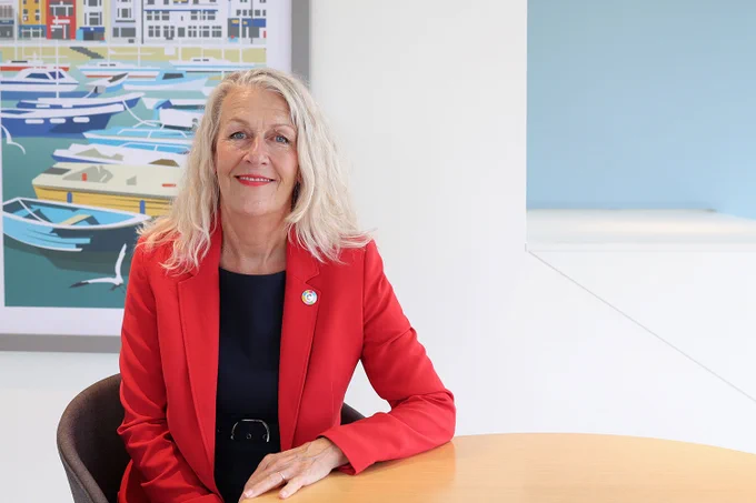 A woman with blonde hair wearing a red blazer smiles at the camera, seated at a table with a harbour artwork behind her.