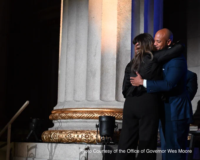 First Lady Dawn Moore is dressed in a dark suit embracing Governor Wes Moore in front of a large stone column with ornate gold detailing. The setting appears to be formal, and the lighting suggests evening or an indoor event.
