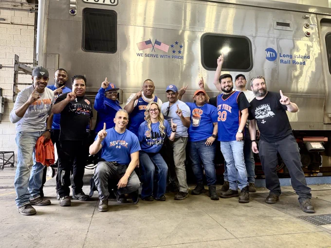 A group of LIRR employees holding one finger up, standing in front of an LIRR M7 train.